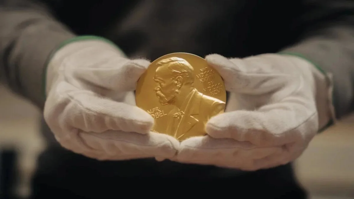 Manos con guantes blancos sostienen una medalla de oro con la efigie de Alfred Nobel.