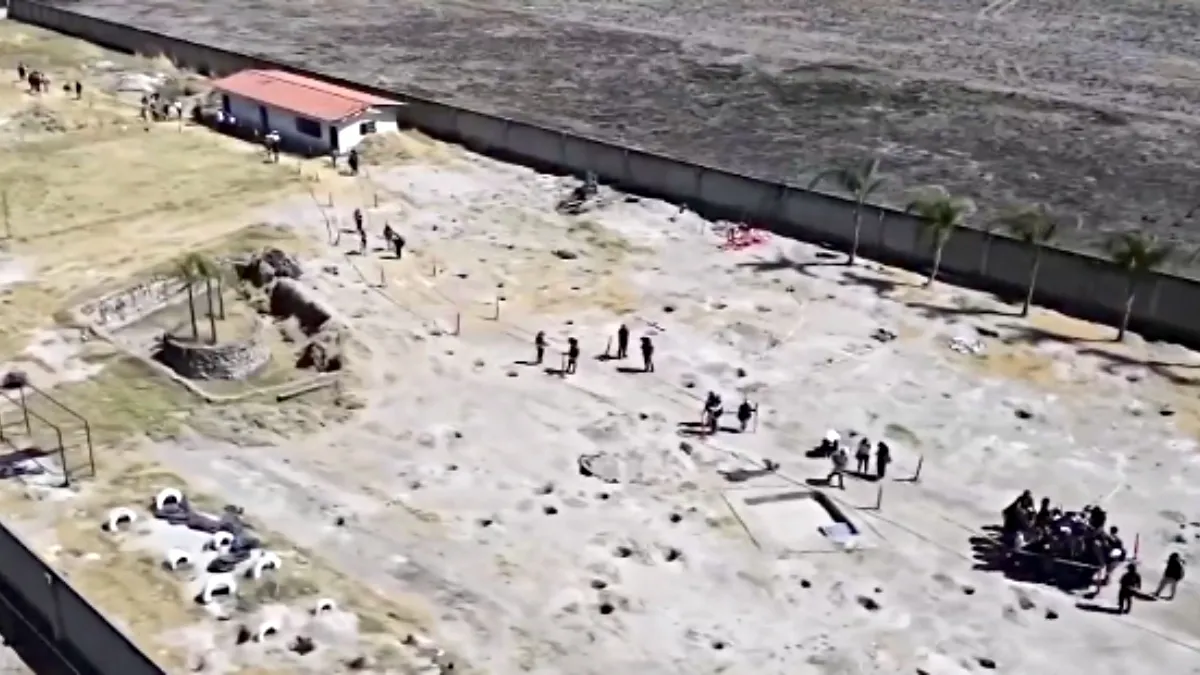 Vista aérea de personas caminando sobre terreno con excavaciones cercanas a una barda y una construcción pequeña.