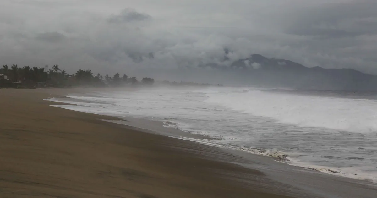 Playa con olas grandes y picadas bajo un cielo nublado y tormentoso con montañas al fondo.