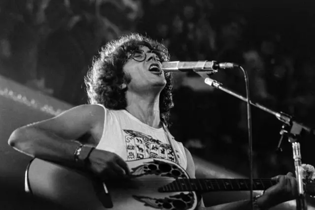 Hombre con anteojos y cabello rizado canta apasionadamente frente a un micrófono mientras toca la guitarra.