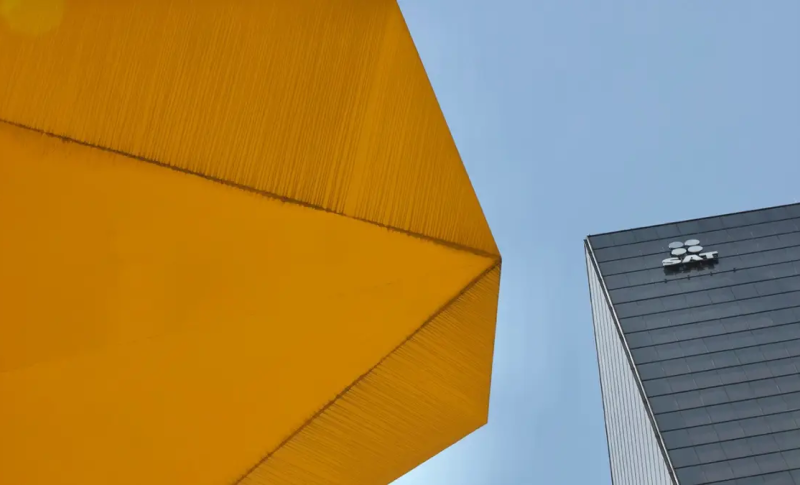 Fotografía de una estructura geométrica amarilla junto a un edificio con el logotipo del SAT bajo cielo despejado.