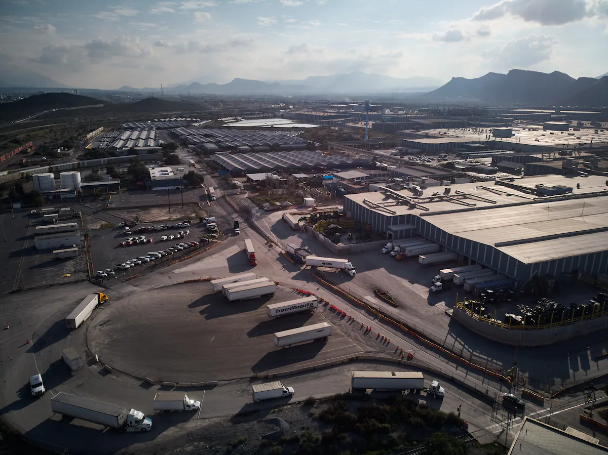 Vista aérea de un complejo industrial con almacenes, camiones de carga y montañas al fondo bajo el sol.