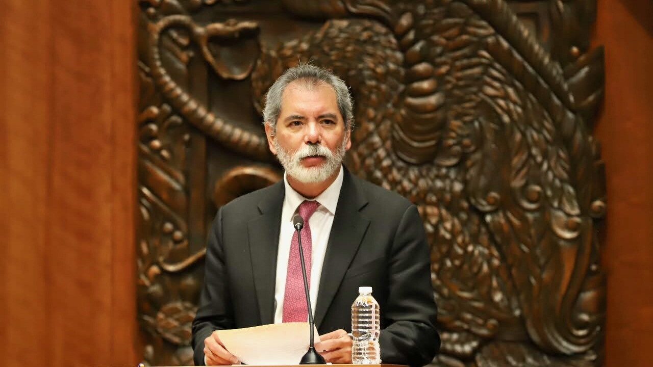 Un hombre de traje y barba hablando en un podio frente a un escudo nacional tallado en madera.