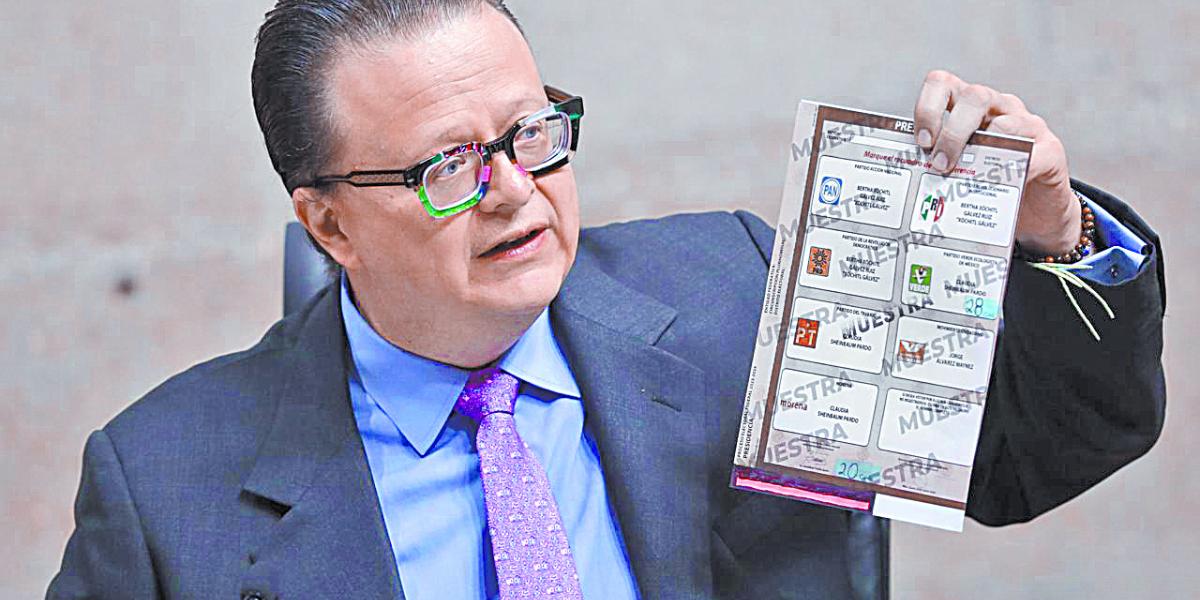 Un hombre con lentes coloridos sostiene una boleta electoral de muestra con logotipos de partidos políticos mexicanos.