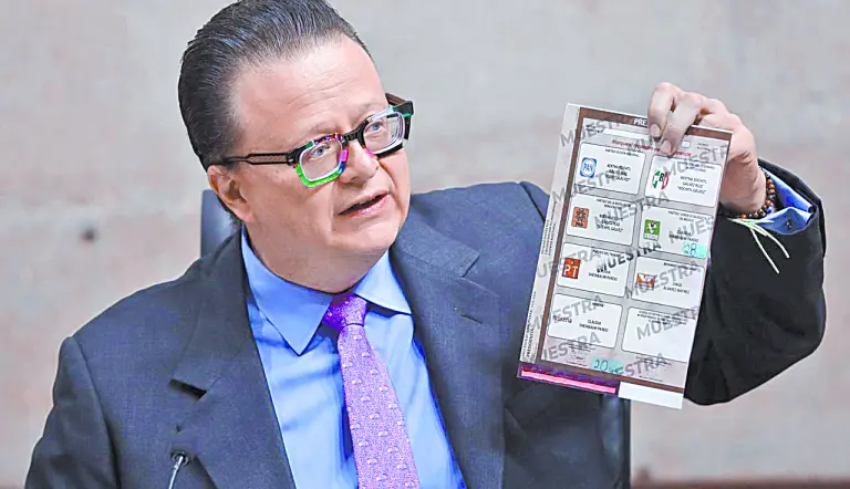 Hombre de traje y lentes sostiene una boleta electoral de muestra con logotipos de partidos políticos mexicanos.